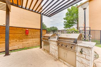 A beige outdoor grill is on a stone pillar under a wooden pergola.